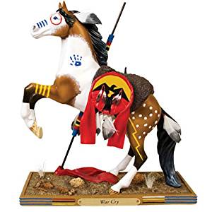 The Trail of Painted Pont collectable War Cry Pony. It shows a rearing paint horse on a desert themed base that has a small plaque that reads War Cry. The horse is a light bay, white, and black horse with a spear on its right side and a has a fur saddle pad with a round decoration attached that shows a flying bird against a sunset sky. The horse has eagle feathers in its mane and tail. The horse also has painted symbols on its body, including a circle around its eye and stripes on its nose.