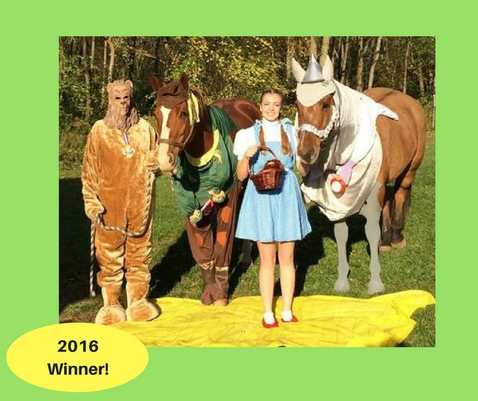 Two people dressed up as Dorothy and the lion from the movie the Wizard of Oz standing with two horses dressed up as the tin man and scarecrow from the same movie.