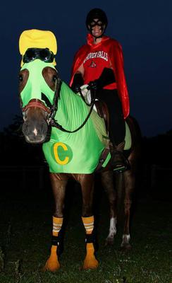 A girl in a red cape riding a horse in a green horse sleezy with a yellow hat and googles on. The horse is also wearing orange socks on its front legs.