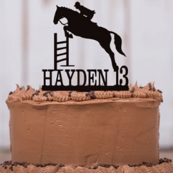The horse and rider jumping cake topper by StrutOurStuff on Etsy. The black cake topper showing a horse and rider jumping a jump above the words Hayden 13 is on a cake with brown frosting.
