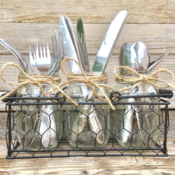 A cutlery caddy by CountryHomeandHeart on Etsy. It is a black metal caddy with three slots and a chicken wire looking outside. There are forks, knives, and spoons tied with burlap in the caddy.