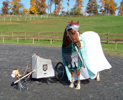A horse dressed up like warrior pyro standing next to a stick horse with a chariot attached to it.