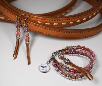 A bracelet and a set of earrings displayed on a light brown set of braided reins. The earrings are made of light brown felt with pink and silver beads. The bracelet is composed of two pieces of brown felt covered in pink and silver beads with a horse charm and a brown felt tassel.