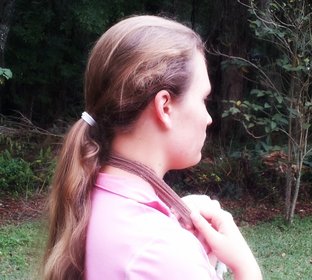 A side view of a girl with waist length brown hair tied back into a ponytail with a white hair tie. The girl is wearing a pink shirt and has a hair netaround her neck and is pulling it forward with her hands. There are plants in the background.
