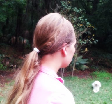 A girl with waist length brown hair tied back into a ponytail with a white hair tie. The girl is wearing a pink shirt and is wearing a hair net like a headband. There is a white dog laying down and plants in the background.