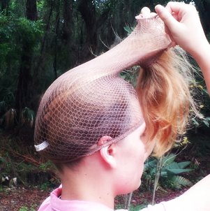 A girl with waist length brown hair tied back into a ponytail with a white hair tie. The girl is wearing a pink shirt and is pulling the hair net up along with the ponytail with both hands. The end of the hair is not in the hair net. There are plants in the background.