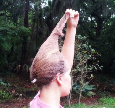 A girl with brown hair tied back into a ponytail with a white hair tie all up in a hair net. The girl is wearing a pink shirt and is using one hand to pull hair net up. All her hair is in the hair net. There are plants in the background