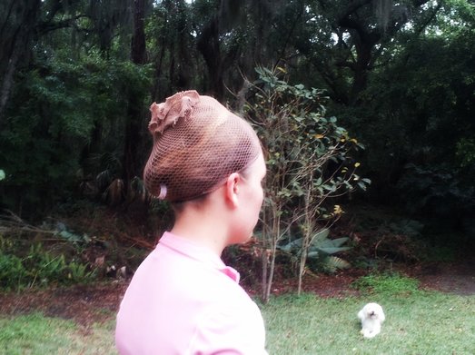 A side view of a girl wearing a pink shirt who has her hair up on top of her head in a hairnet. There is a white dog facing the camera, and plants in the background.