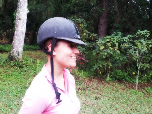 A side view of a girl in a pink shirt smiling. Her hair is up in a GPA riding helmet. There are plants in the background.