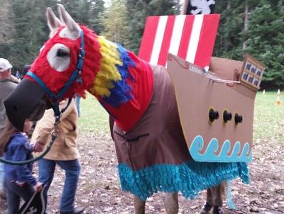 A horse with a neck cover that looks like parrot feathers and a pirate ship made of cardboard covering the horse's body.