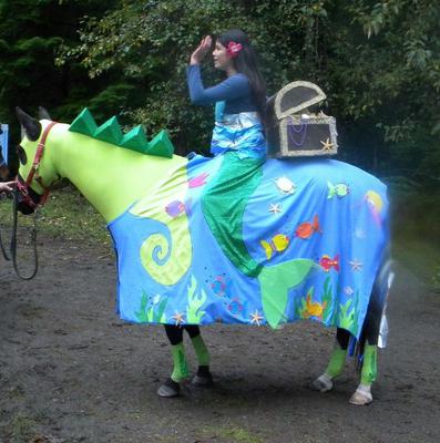 A horse dressed up like a seahorse with a girl dressed like a mermaid riding the horse.