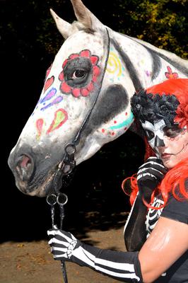 A girl in a skeleton costume standing beside a horse with different drawings such as a flower around its eye, different color dots, and colorful stripes on its body.