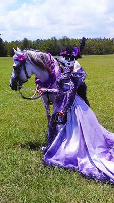 A woman dressed up as a dead bride in a purple dress standing next to her horse who is decorated with purple accessories.