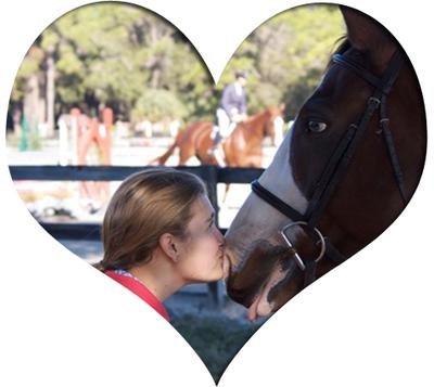 A picture of a girl kissing a horse on the muzzle. The horse has a blue eye, a white blaze, is wearing an english bridle, and is sticking its tongue out. The girl has brown/blonde hair and is wearing a pink top. There is a horse and rider along with jumps in the background. There is a heart frame around the image.