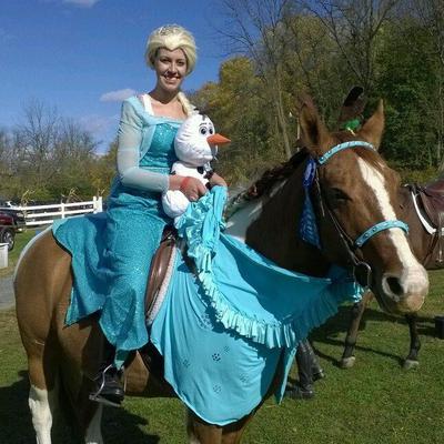 A girl dressed like Elsa from the movie Frozen holding a stuffed animal replication of Olaf from the movie Frozen. She and the stuffed animal are on a horse dressed in blue.