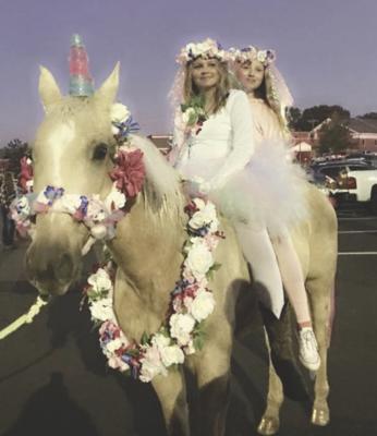 A horse dressed up as a unicorn with a flower halter and a ring of flowers hanging on her neck. Two girls dressed as fairies are sitting on the horse.