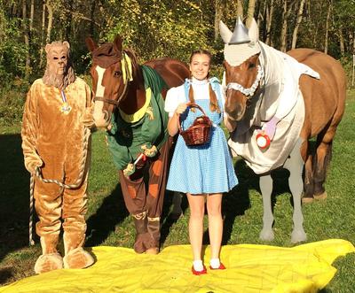 Two horses one dressed up as the scarecrow another as the tin man standing beside two people one dressed as the lion another dressed as Dorothy from the movie The Wizard of Oz.