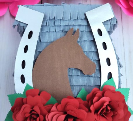A pinata that has a dark gray background with a silver horseshoe on top, a brown horse head silhouette, and three red flowers on the bottom.