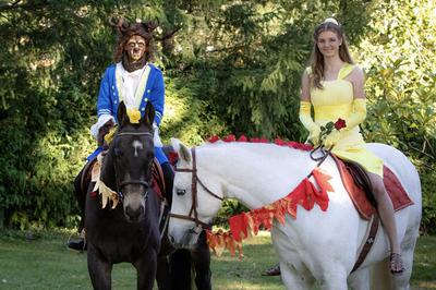 A girl dressed up like Belle on a white horse and a guy dressed up like the beast from beauty and the beast on a black horse.