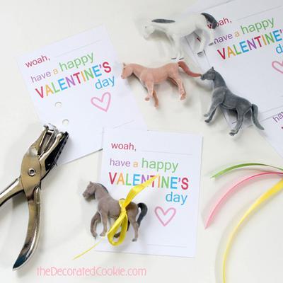 A photo from TheDecoratedCookie.com that shows valentine cards with words woah, have a happy valentine's day in rainbow colored font. There are four horse figurines, one a chestnut, another a bay, another a black horse, and the fourth a grey horse. There is also a hole punch on the left side and ribbon on the right. 