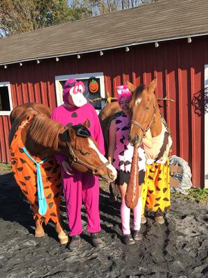 Two people dressed up like dinosaurs standing next to two horses dressed up like Flintstone characters.