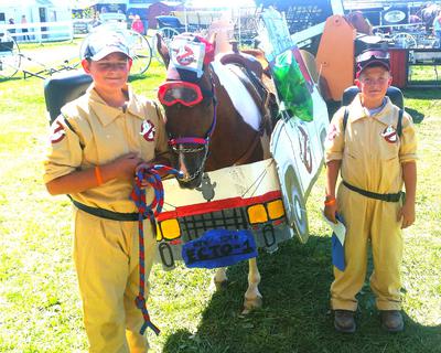 A horse dressed up like the ghostbusters van standing next to two boys dressed up like ghostbusters.