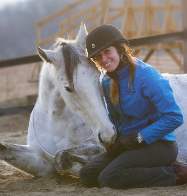 Image from Harmony Horsemanship Academy featuring a woman next to a dapple grey horse laying down