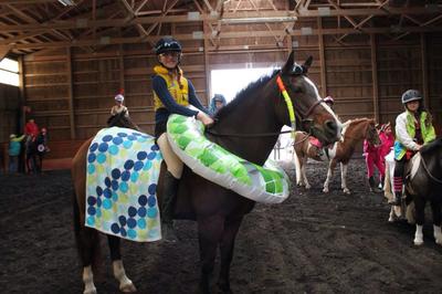 A horse with a pool floaty around his/her neck and a pool blanket over her butt. The rider is wearing a life jacket.