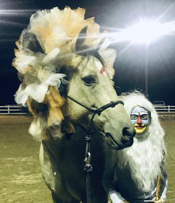 A buckskin horse with a fake colorful looking lion mane on is standing next to a girl wearing a white wig with face paint on that looks like a monkey.
