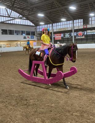 The horse is wearing two cutouts that look like the bottom of a rocking horse. A girl dressed up like a little kid is riding the horse.