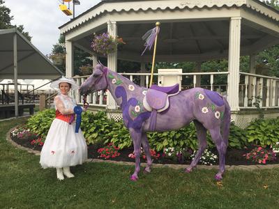 A girl wearing a Mary Poppins outfit standing next to a horse that is painted in purple with flowers and a pole attached to the saddle to make the horse look like a carousel horse.