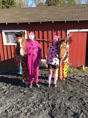 Two people dressed up as dinosaurs are standing beside two horses each dressed up as a Flintstone character.